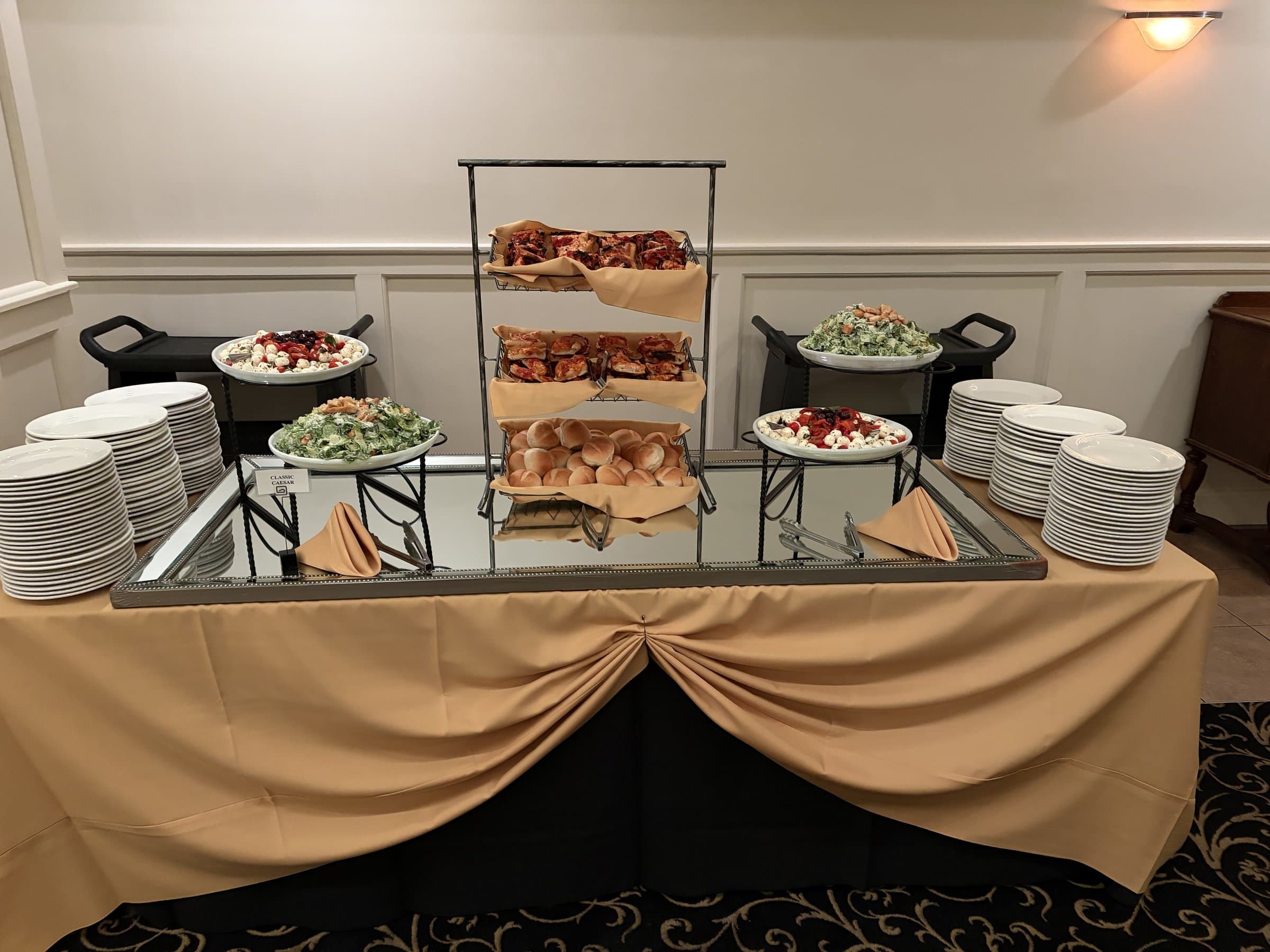 A skirted buffet station with a Classic Caesar placard — tiered baskets of focaccia and dinner rolls, bowls of Caesar and caprese salad, and stacks of white plates